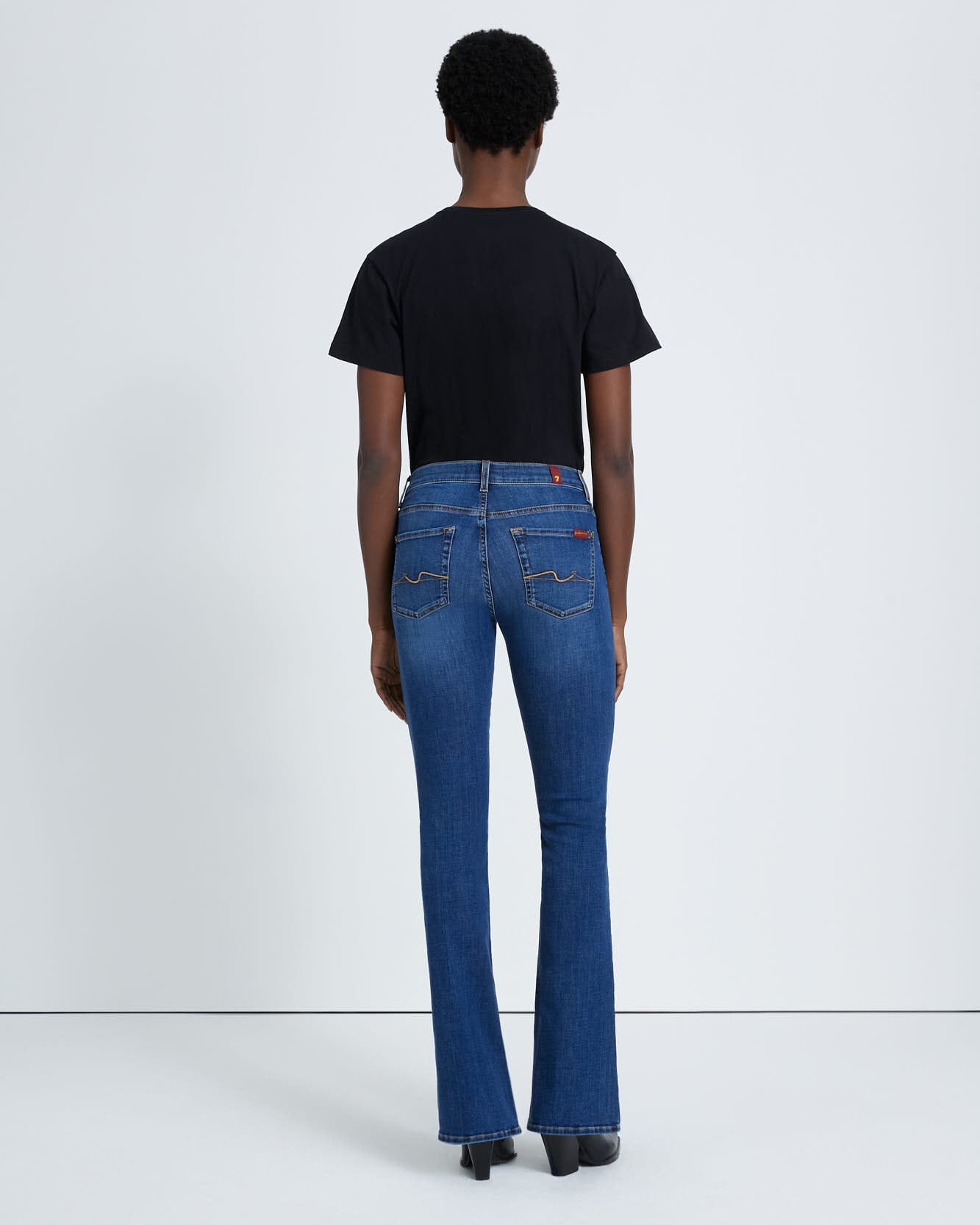 model wearing 7 for all mankind kimmie mid rise boot cut jean in duchess blue wash, paired with a black t-shirt and black boots, shown from the back