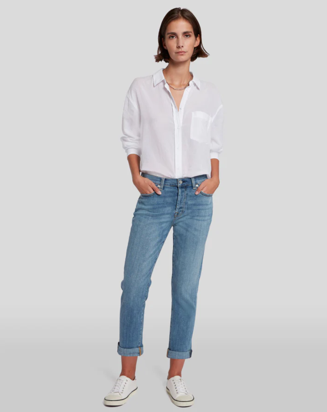 model wearing 7 for all mankind josefina boyfriend fit jean in bright light broken twill blue, paired with a white blouse and white sneakers, posed with hands in pockets